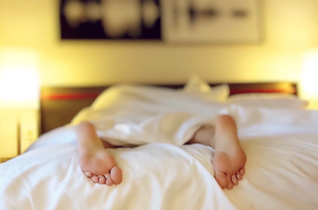 Človek s odkrytými nohami leží na posteli zakrytý dekou 