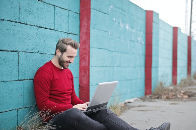 Muž v červenom svetri sedí pri tyrkysovom múre píše na notebooku